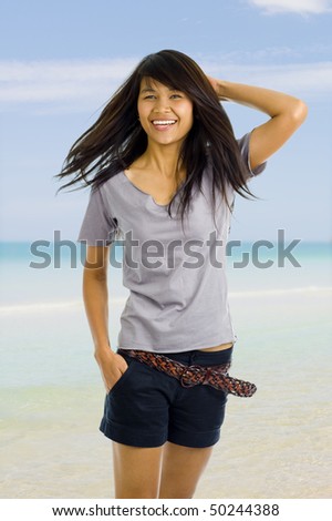 beautiful, young asian woman with one hand in her short\'s pocket and the other one behind her head, posing in front of a beach