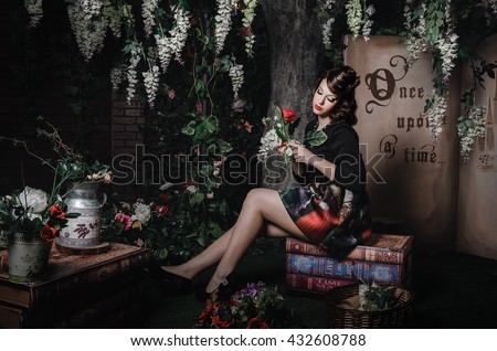 Fashion portrait of romantic beautiful girl with hairstyle, red lips, art dress, holding rose flower, sitting on books. Princess in mistery forest. Creative concept Once upon a time in fantasy.