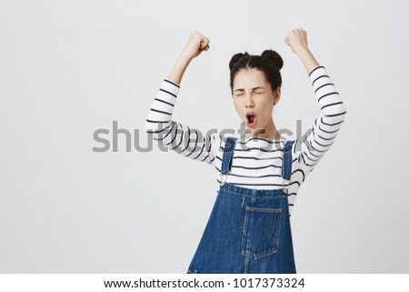 Brunette girl with hairbuns in striped top excited and glad to achieve victory, clenches fists, screams in excitement with closed eyes, happy to pass all exams at university, successful person.
