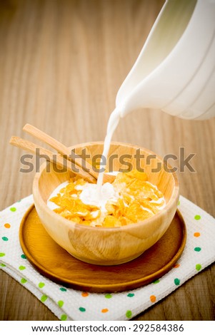 Milk pouring into a wooden bowl of nutritious and delicious corn flake cereal on wooden table. Darken effect.