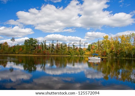 Photograph of a quiet and remote northwoods lake on a beautiful autumn day with clouds reflected in the still blue waters while a pontoon boat of sightseers slowly motors by.