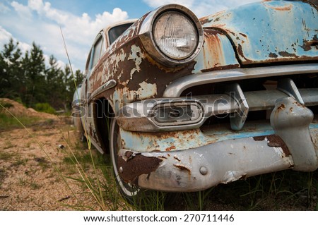 Over Parked. Rusted, classic car sits broken down in a sandy lot with trees and clouds in the distance.