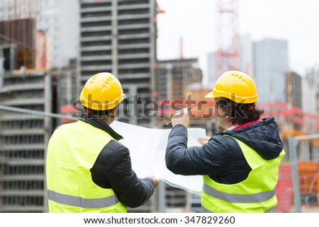 Back turned architect and technician at construction site reviewing office blueprints