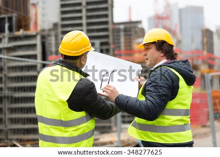 Construction worker with yellow hardhat and safety jacket checking blueprint with an architect