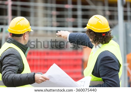 Back turned construction workers with yellow hardhat and safety jacket checking office blueprint