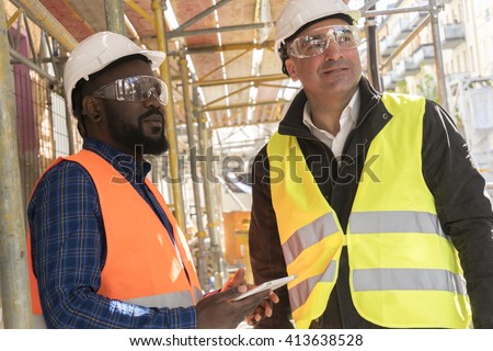Two construction workers, a black and a white, wearing orange and yellow safety jackets and helmets among scaffolding on construction site
