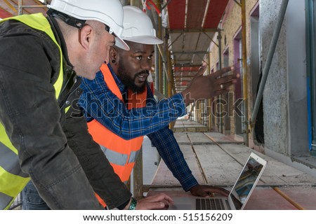 Two construction workers, an African american and a white, wearing orange and yellow safety jackets and helmets among scaffolding on construction site