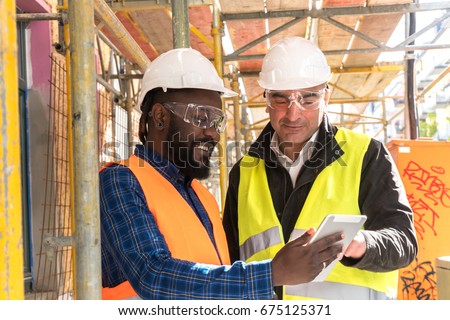 Two engineers, an African American and a caucasian, wearing orange and yellow safety jackets and helmets, working with tablet computer among scaffolding on construction site