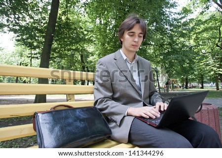 Smart businessman sitting on a wooden bench in a city park and using his laptop computer, outdoors.