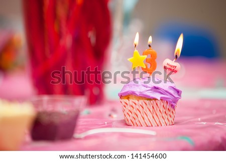 A close up of a birthday purple icing cupcake with a star, number three and a happy birthday candles all lit.