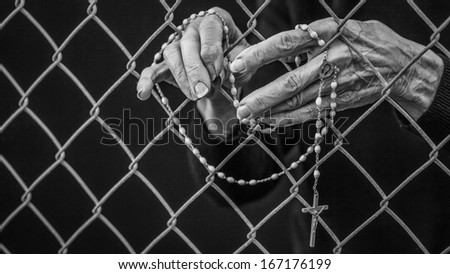 A close up of an old woman\'s hand holding her rosary through a chain link fence.