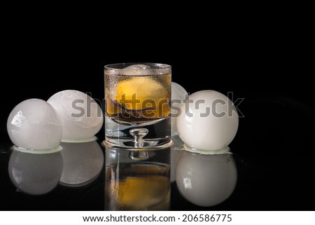 A studio shot of a low-ball glass of whiskey with an ice ball in the glass and surrounded by ice  balls with a black background.