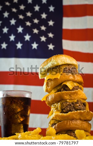A close up of a five patty cheese burger, fries, iced soda and US flag in the background.