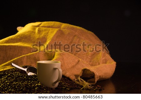 A close up of a burlap coffee bag opened with coffee beans spilling out with a cup of coffee in the foreground.