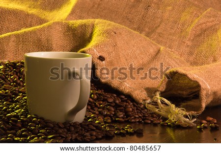 A close up of a burlap coffee bag opened with coffee beans spilling out with a cup of coffee in the foreground.