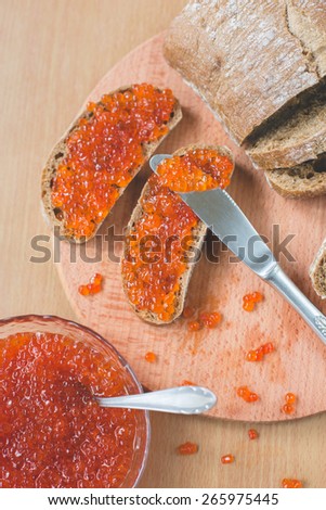 Red caviar toasts sandwich with silver knife, bowl of tasty red caviar with spoon and fresh baked homemade healthy bread on a wooden table background