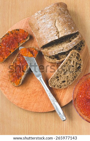 Red caviar toasts sandwich with silver knife, bowl of tasty red caviar with spoon and fresh baked homemade healthy bread on a wooden table background