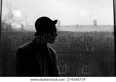 portrait of a handsome caucasian guy in hat silhouette on a rain drops on window and city view background