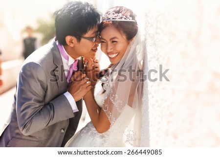 happy stylish asian bride and groom smiling and holding hands  near wall of island Santorini