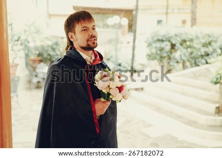 Stylish elegant beard rich groom in the mantle with bouquet of flowers roses peonies background walls stairs