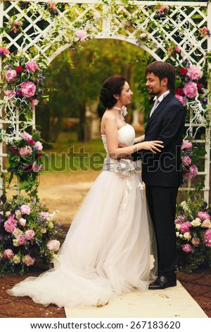 wonderful stylish rich happy bride and groom smiling at a wedding ceremony holding hands look at each other in green garden near white arch with flowers Rome Italy