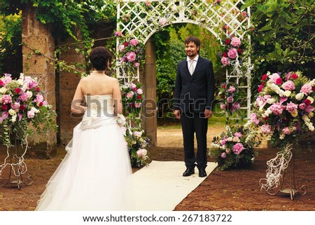 wonderful stylish rich happy bride and groom at a wedding ceremony look at each other in green garden near white arch with flowers Rome Italy