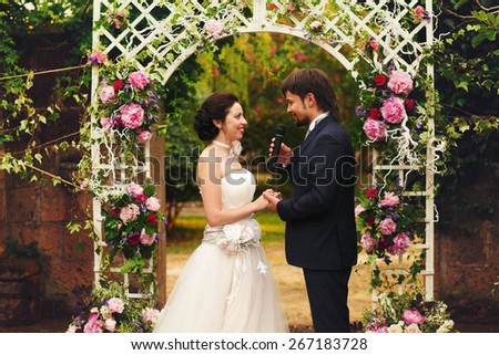 wonderful stylish rich happy bride and groom holding hands at a wedding ceremony look at each other in green garden near white arch with flowers Rome Italy