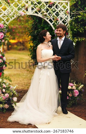 wonderful stylish rich happy bride and groom smiling at a wedding ceremony holding hands look at each other in green garden near white arch with flowers Rome Italy