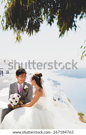 happy stylish asian bride and groom look sitting on a wall roof of a white house under green tree on street of island Santorini background sky and sea
