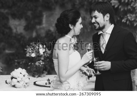 rich stylish happy bride and groom kissing smiling holding goblets with champagne  near a white wedding table decorated with flowers peonies and candles Rome Italy black and white