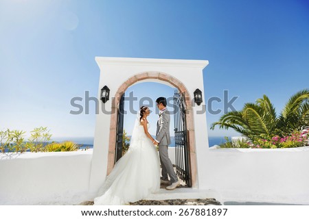 happy stylish asian bride and groom look at each other and holding hands  under arch  on background blue sky and sea in island Santorini, Greece