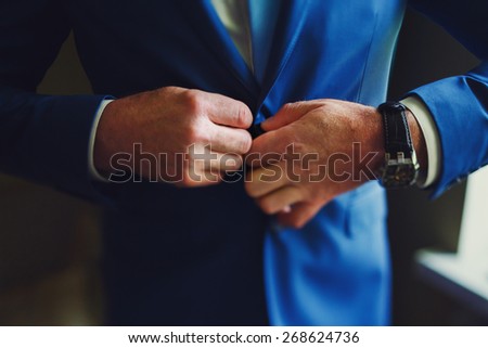 adult solid groom button his blue suit near window