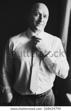 solid bald man in shirt standing near the window straightens tie looks straight black and white