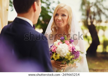wonderful stylish rich happy bride and groom holding hands look et each other at a wedding ceremony in  garden near arch with flowers