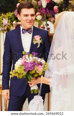 wonderful stylish rich happy bride and groom holding hands look et each other at a wedding ceremony in green garden near white arch with flowers