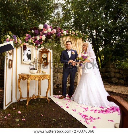 wonderful stylish rich happy bride and groom holding hands at a wedding ceremony in  garden near arch with flowers