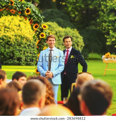 rich stylish groom with bestman waiting bride near the arch of sunflowers
