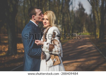 elegant stylish groom hugging and kissing gorgeous blonde bride on the background of trees in the park