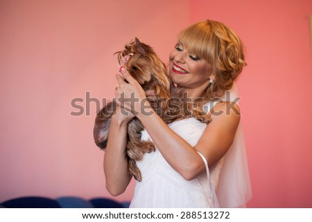 luxurious stylish smiling blonde bride with red lips is holding yorkshire terrier on the background pink wall