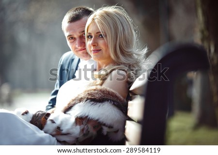elegant groom hugging his stylish happy blonde bride and smiling on the background of park sitting on the bench