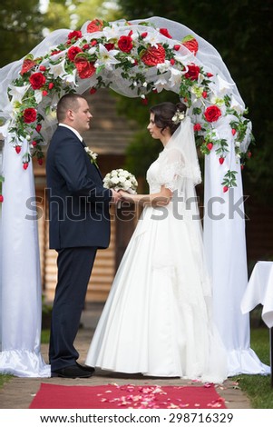 stylish wedding of happy couple under elegantly decorated with red roses and white lilies arch