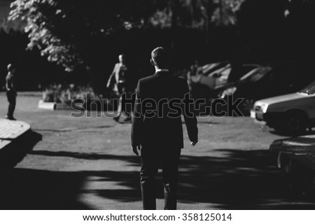 Man in business suit walks on the road against the background of the city park and parked cars. Black and white portrait of man from the back.