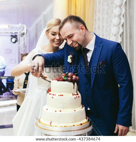 Groom looks curious trying to cut a wedding cake together with a bride