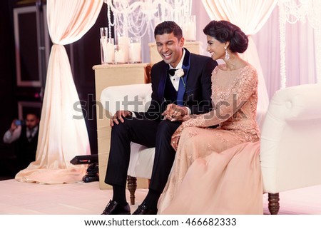 Laughing groom holds a mehndi hand of Indian bride while sitting on the white sofa during the wedding dinner