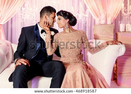 Glorious Indian woman holds face of handsome brunette man while sitting on white sofa in pink room