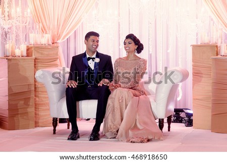 Smiles of stunning Indian wedding couple shine while they sit on the cosy sofa in a luxury hall
