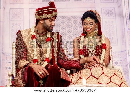 Pretty Indian bride in red suit smiles while groom touches her delicate hands