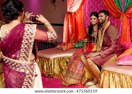 Woman in saree makes picture of smiling Indian wedding couple sitting on the sofa with silk pillows