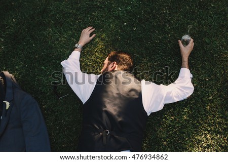 Look from above at a drunk man in black waistcoat lying on the grass with bottle of whisky