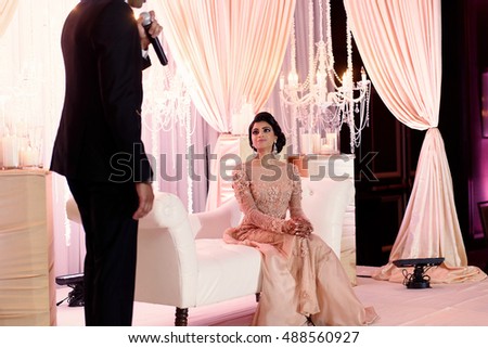 Adorable Indian bride sits on the white sofa in pink gown while groom tells her something in the microphone